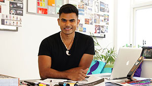 Jason Fenandes smiling at the camera with his arms folded and resting on a desk with a MacBook laptop