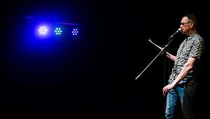 Rick Sanders on a darkened stage with blue lights, speaking into a microphone