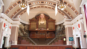 A large organ at the top of a flight of wooden stairs in a tall, ornate hall with arching roof supports